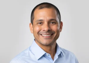 Professional headshot of male dentist smiling wearing light blue shirt against neutral background.